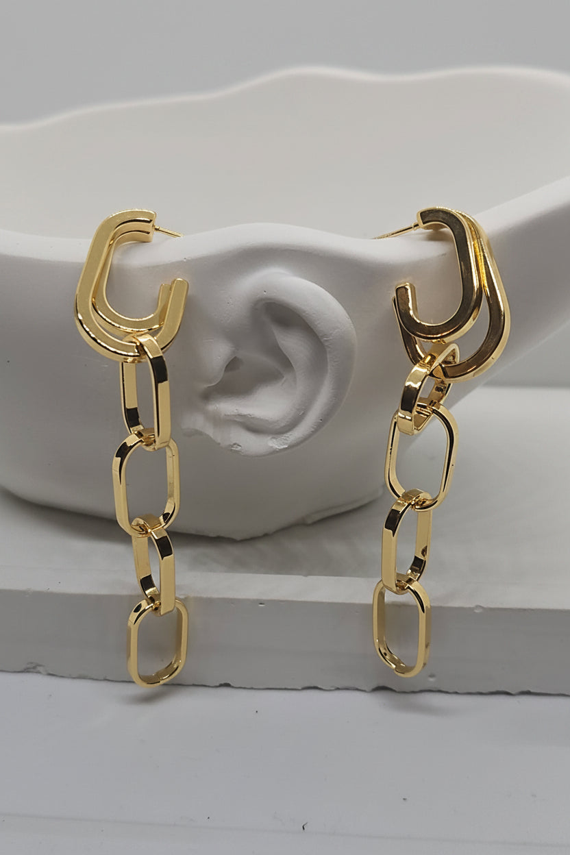 A pair of gold-toned hoop earrings with dangling chain-link drops, displayed on a white sculptural ear-shaped stand against a light gray background.