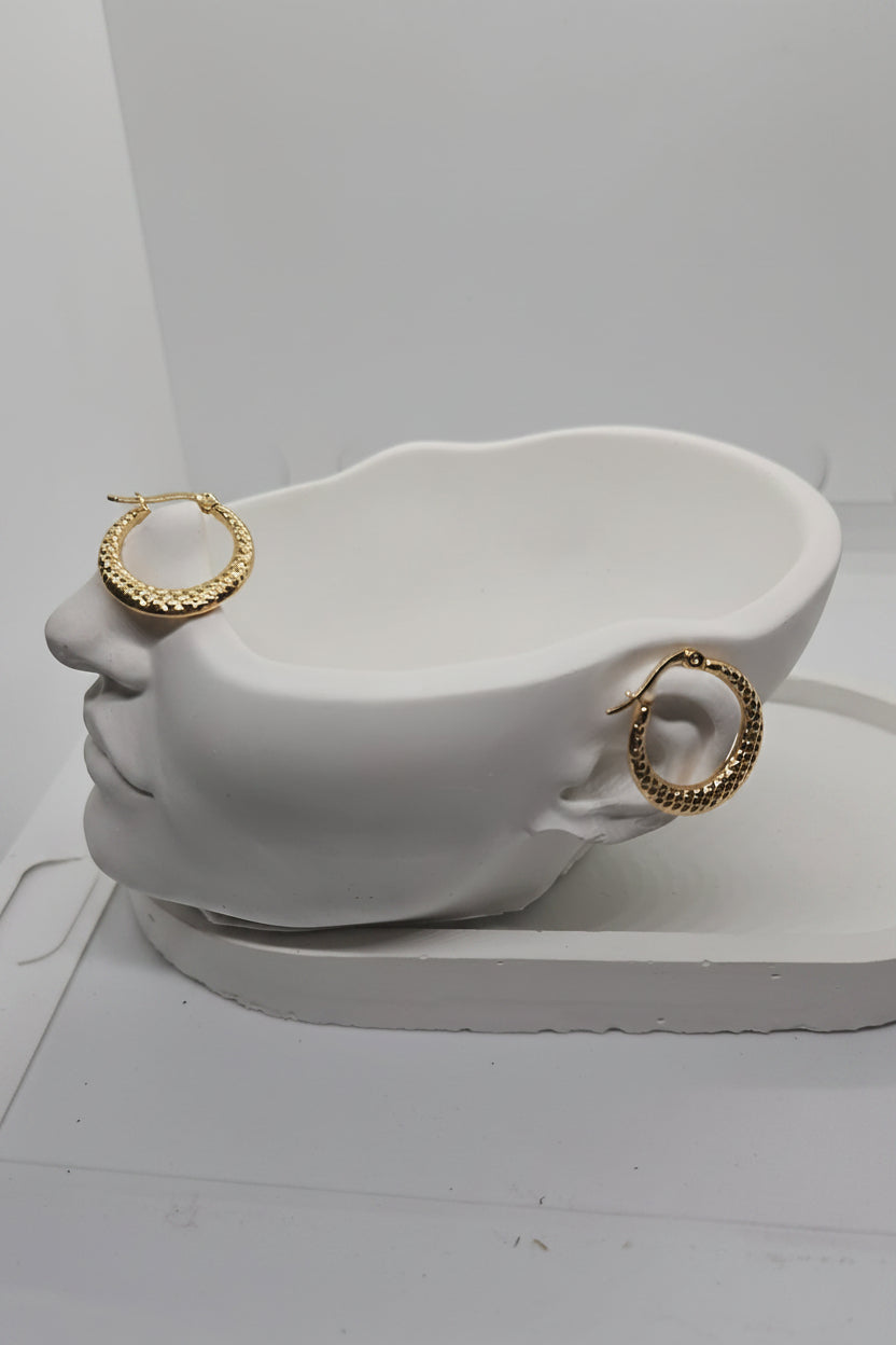 A pair of textured gold hoop earrings displayed on a white sculptural head-shaped jewelry stand against a plain white background.