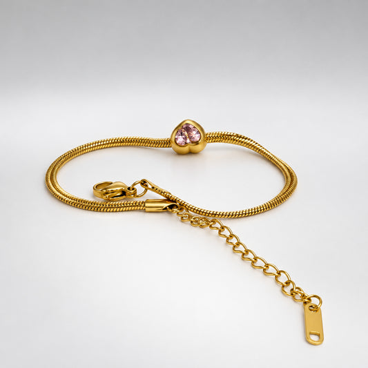 A gold-colored snake chain bracelet with a small pink heart-shaped crystal charm and an adjustable lobster clasp with a chain extender, displayed on a white surface.
