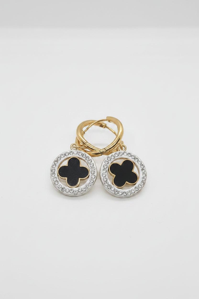 A pair of gold and silver hoop earrings with black clover motifs, displayed on a textured white surface.