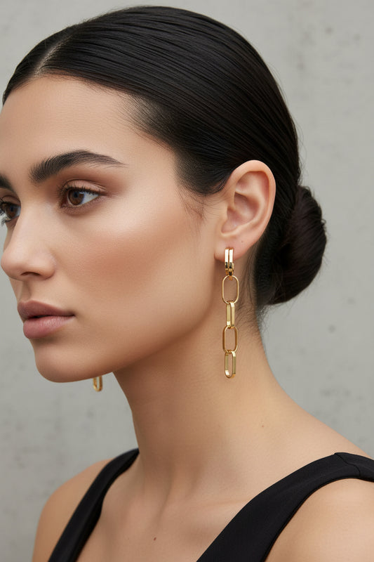 A pair of gold huggie hoop earrings with elongated link details, displayed on a white ceramic dish against a light gray background.