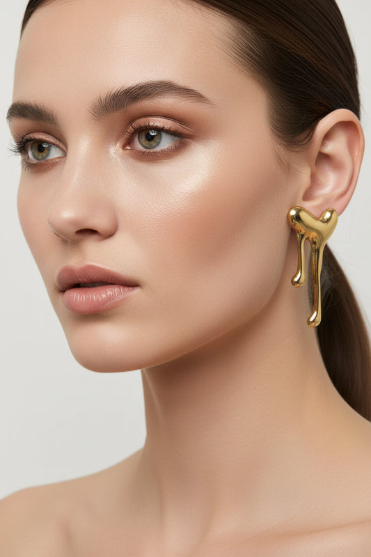 A pair of gold-toned, abstract sculptural stud earrings with a dripping or melting organic shape, displayed against a neutral light gray background.