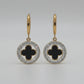 A pair of gold hoop earrings with circular drop charms featuring a black clover motif surrounded by a silver-tone halo of small clear stones, displayed on a textured white surface.