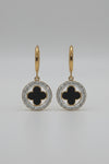 A pair of gold hoop earrings with circular drop charms featuring a black clover motif surrounded by a silver-tone halo of small clear stones, displayed on a textured white surface.