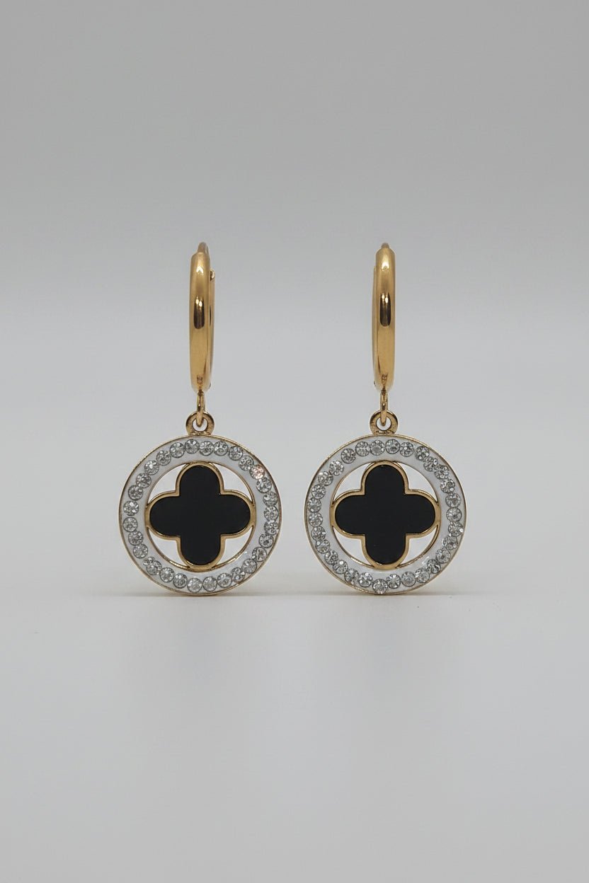 A pair of gold hoop earrings with circular drop charms featuring a black clover motif surrounded by a silver-tone halo of small clear stones, displayed on a textured white surface.