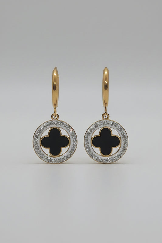 A pair of gold hoop earrings with circular drop charms featuring a black clover motif surrounded by a silver-tone halo of small clear stones, displayed on a textured white surface.