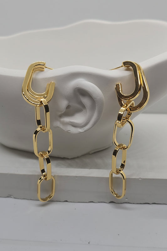 A pair of gold-toned hoop earrings with dangling chain-link drops, displayed on a white sculptural ear-shaped stand against a light gray background.