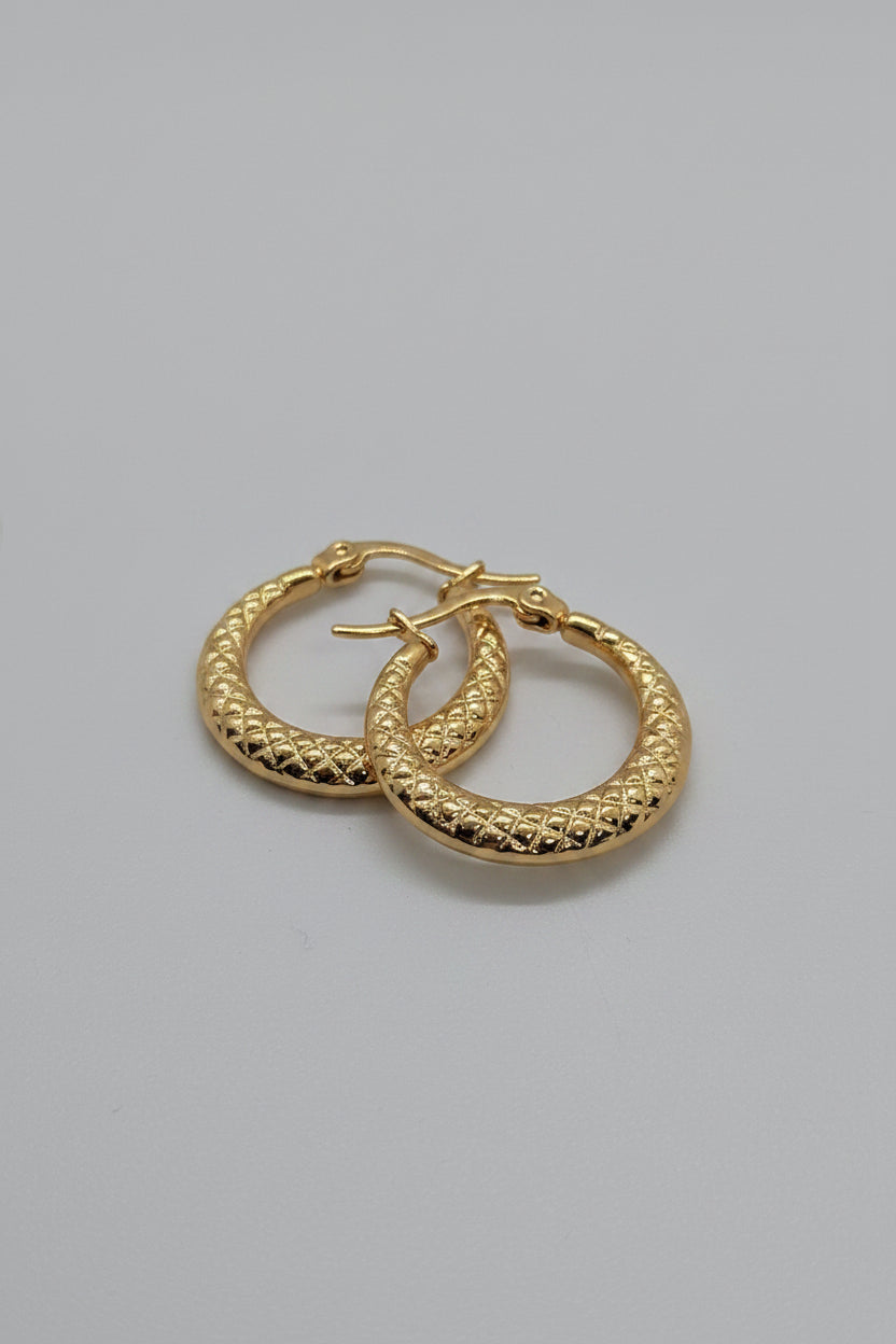 A pair of textured gold hoop earrings with a diamond-patterned beaded surface, displayed on a white textured stone surface.