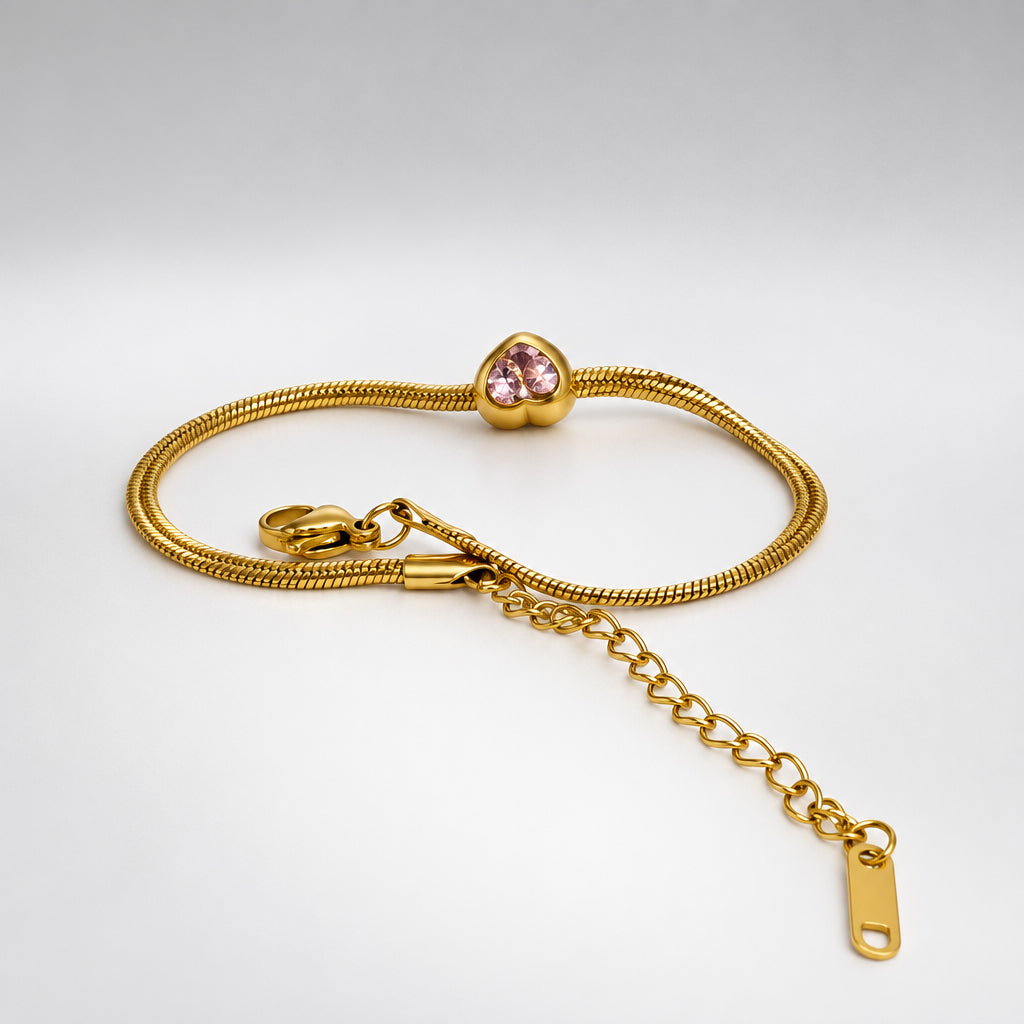 A gold-colored snake chain bracelet with a small pink heart-shaped crystal charm and an adjustable lobster clasp with a chain extender, displayed on a white surface.