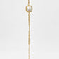 A gold chain bracelet featuring a central square bezel-set white pearl and a dangling chain with a small round gold bead at the end, displayed against a light beige background.