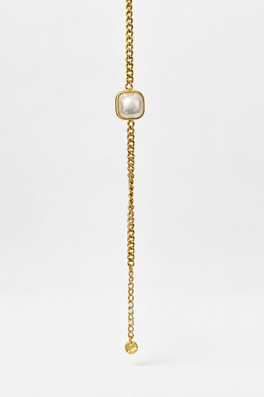 A gold chain bracelet featuring a central square bezel-set white pearl and a dangling chain with a small round gold bead at the end, displayed against a light beige background.