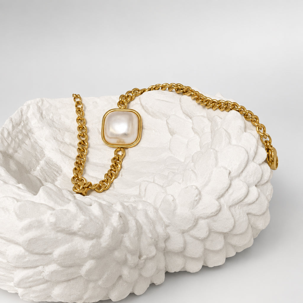 A gold chain necklace with a square pendant featuring a white faux pearl, displayed on a textured white ceramic dish against a neutral beige background.