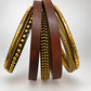 A set of vintage bangles or wristlets hanging from a thin wire stand against a plain light background. The set includes three dark brown wooden pieces with rows of small metallic beads, two gold-toned metal pieces with beaded detailing, and two gold-toned pieces with a textured, ribbed outer surface.