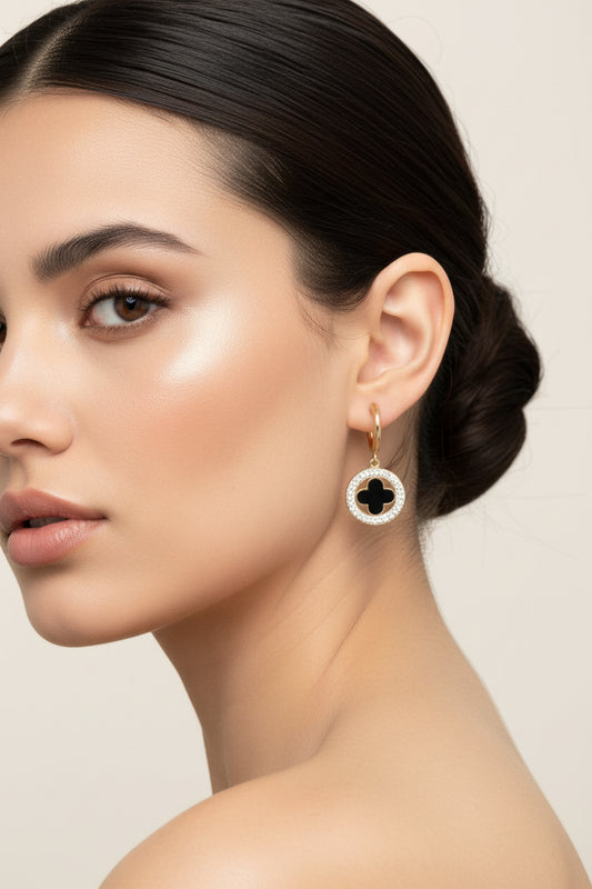 A pair of gold hoop earrings with oval dangles featuring a black clover motif surrounded by clear rhinestones, displayed on a white surface.
