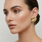 A pair of gold-toned textured stud earrings with a ribbed, segmented design and a smooth curved top accent, displayed on a light gray surface.