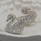 A pair of silver-tone swan-shaped earrings covered in clear rhinestones, displayed on a textured white stone surface.