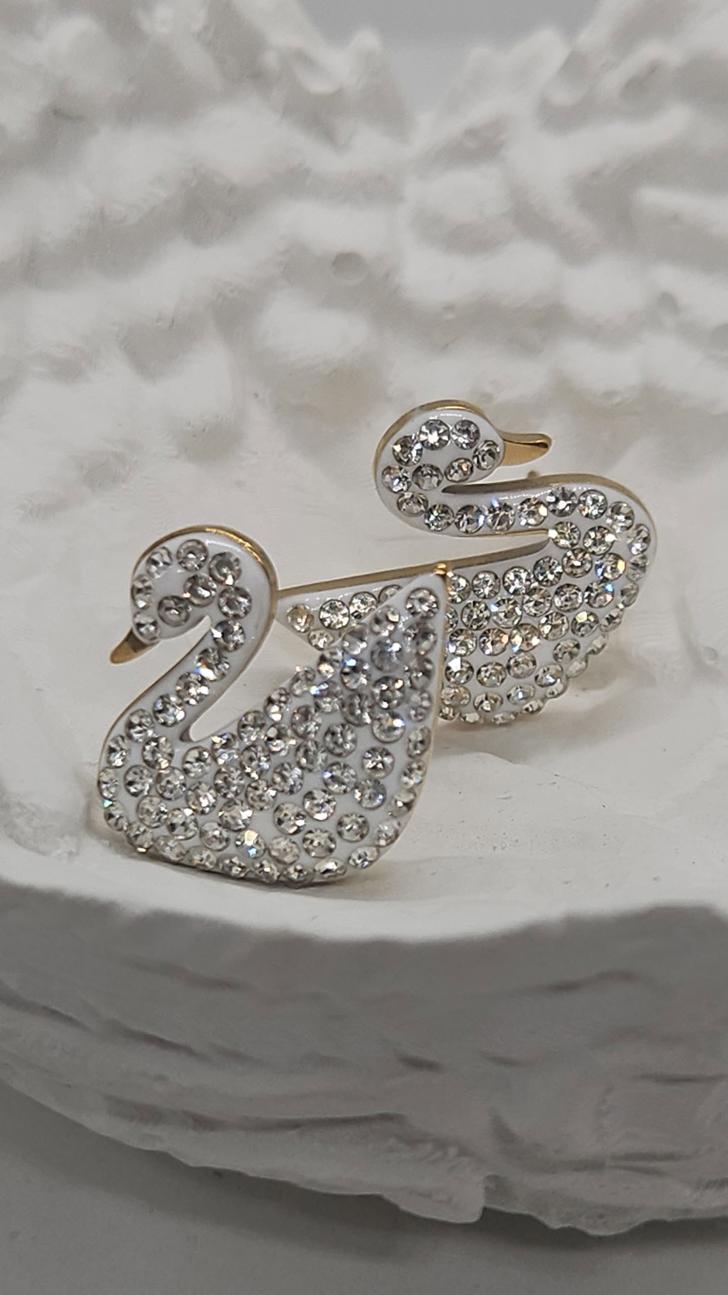 A pair of silver-tone swan-shaped earrings covered in clear rhinestones, displayed on a textured white stone surface.