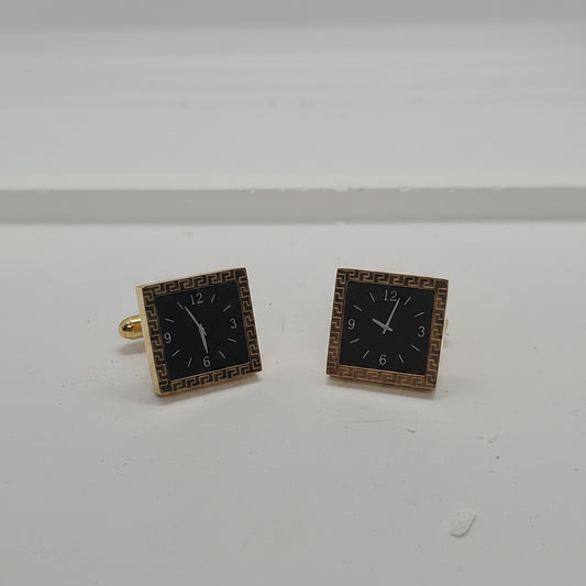 A pair of square earrings designed to look like clock faces, featuring black centers with white numerals and hands, surrounded by gold-tone borders.