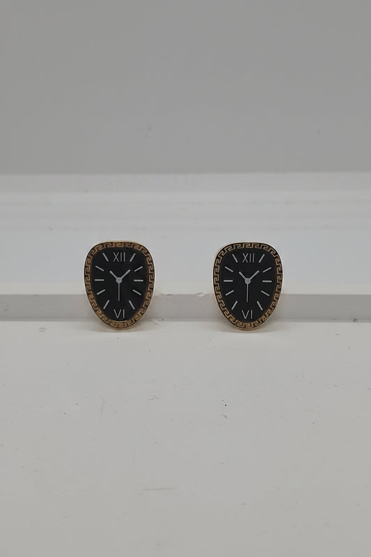 A pair of vintage-style clock face stud earrings with black enamel centers, gold-tone Roman numerals, and gold-tone bezels, displayed on a white surface against a light gray background.