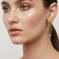 A pair of gold-toned, abstract sculptural stud earrings with a dripping or melting organic shape, displayed against a neutral light gray background.