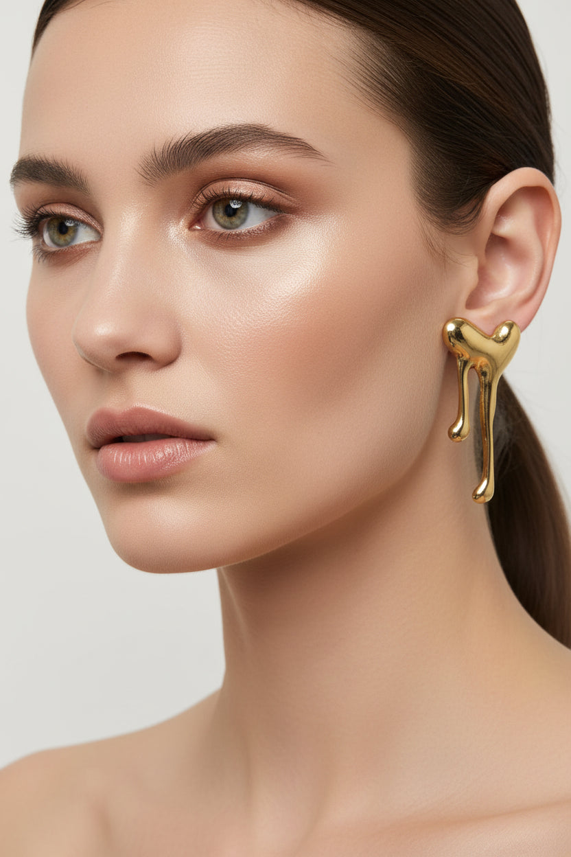 A pair of gold-toned, abstract sculptural stud earrings with a dripping or melting organic shape, displayed against a neutral light gray background.